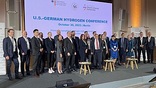 A big group of people in business attire standing on a stage with roll-ups and a power point presentation in the background.