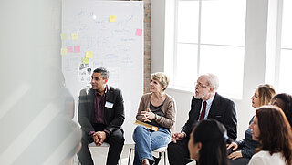 People of different genders, ages and skin colours sit in a circle of chairs and discuss. In the background is a whiteboard with notes and post-its.