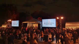 Outdoor evening event with several people standing in small groups talking. Big screens with presentations in the background.