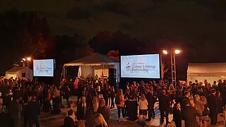 Outdoor evening event with several people standing in small groups talking. Big screens with presentations in the background.