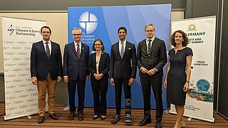 Two women and four men in business attire standing in front of roll-ups smiling. 