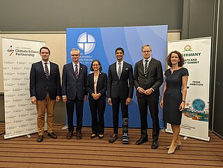 Two women and four men in business attire standing in front of roll-ups smiling. 