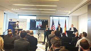 Several people sitting on rows of chairs at a conference venue. Four people are standing at the front behind high tables, screens with a presentation as well as the U.S. and German national flags in the background.