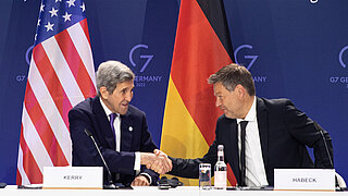 Two men are sitting at a desk shaking hands. The national flags of Germany and the U.S. are in the background.