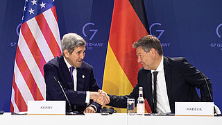 Two men are sitting at a desk shaking hands. The national flags of Germany and the U.S. are in the background.