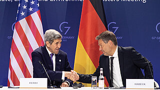 Two men are sitting at a desk shaking hands. The national flags of Germany and the U.S. are in the background.