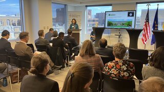 Several people sitting on rows of chairs at a conference venue. The U.S. and European flags are placed on a stage where a person is giving a presentation. A screen with presentation slides is in the background.